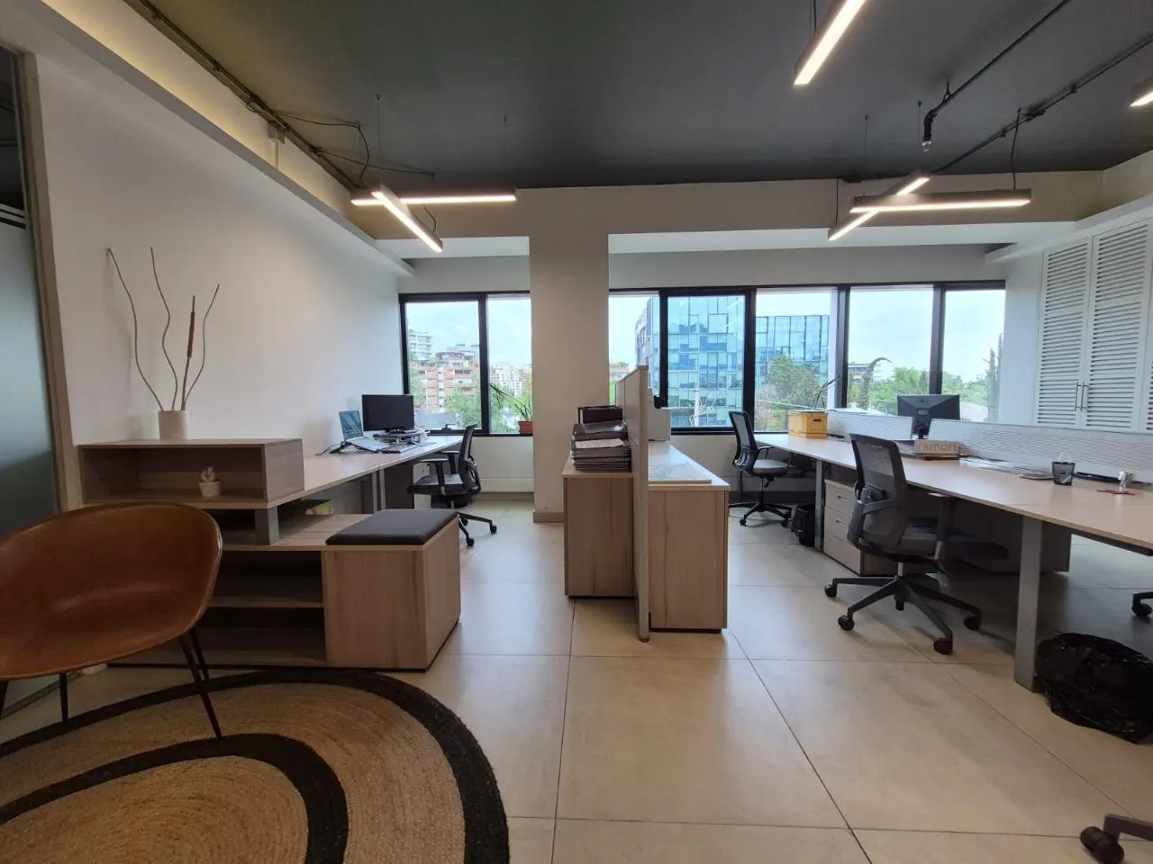 Bright office space with desks, chairs, and computers. Large windows offer city views. A brown chair and rug add warmth to the modern interior.