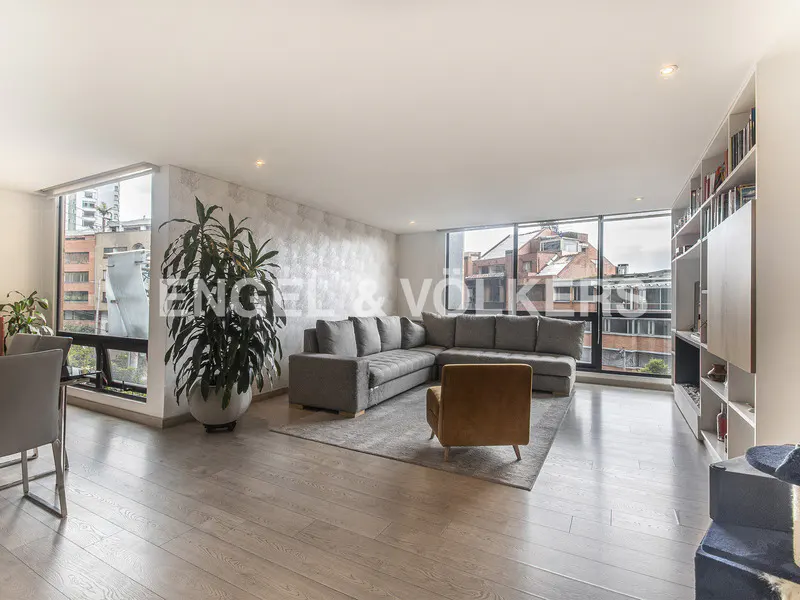 Bright living room with a gray sectional sofa, brown chair, wood floors, and large windows with city views.