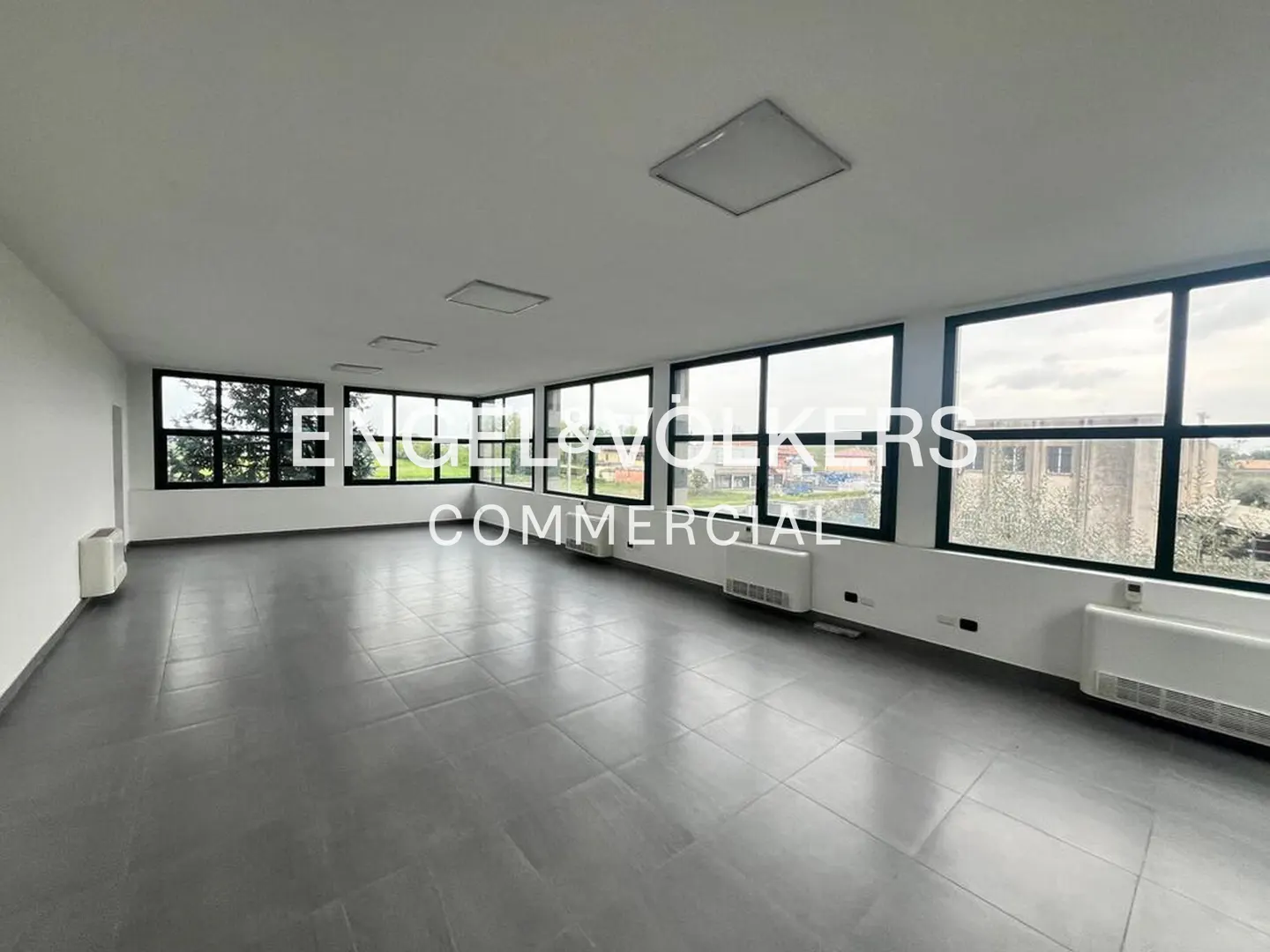 Bright, empty commercial space with gray tile floor, white walls, and large windows with black frames. Radiators line the walls.