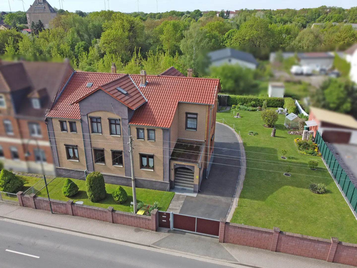 Aerial view of a two-story tan house with a red tile roof, a paved driveway, and a large green backyard with a brick fence.