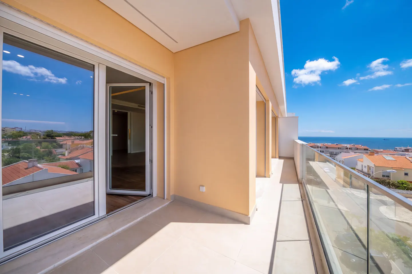 Balcony view with glass railing, beige walls, and open sliding glass doors. Ocean and rooftops visible in the background under a blue sky.