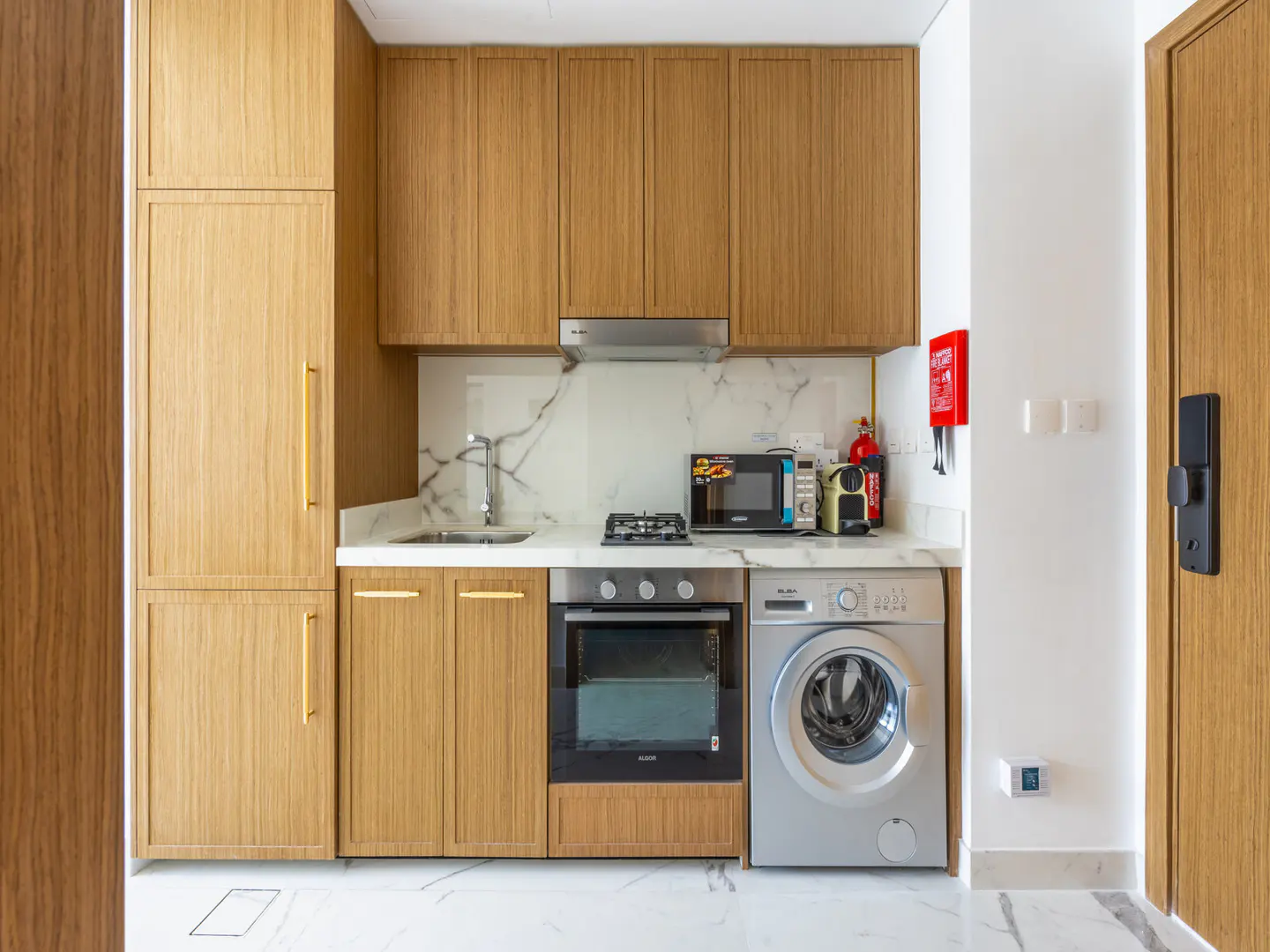 A compact kitchen features wood cabinets, marble countertops, a sink, oven, microwave, and a washing machine. A red fire blanket hangs on the white wall.