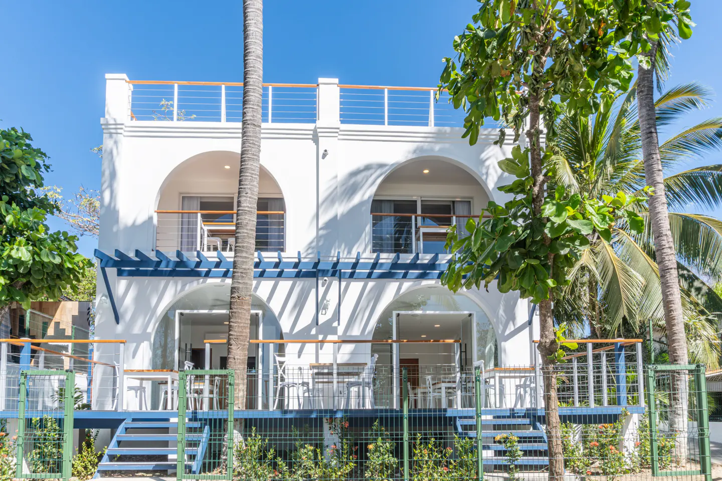 Exterior of a white, two-story building with arched doorways and blue accents, surrounded by tropical trees.