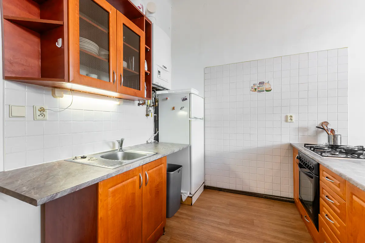 A kitchen with wood cabinets, a stainless steel sink, a white refrigerator, and white tiled walls.