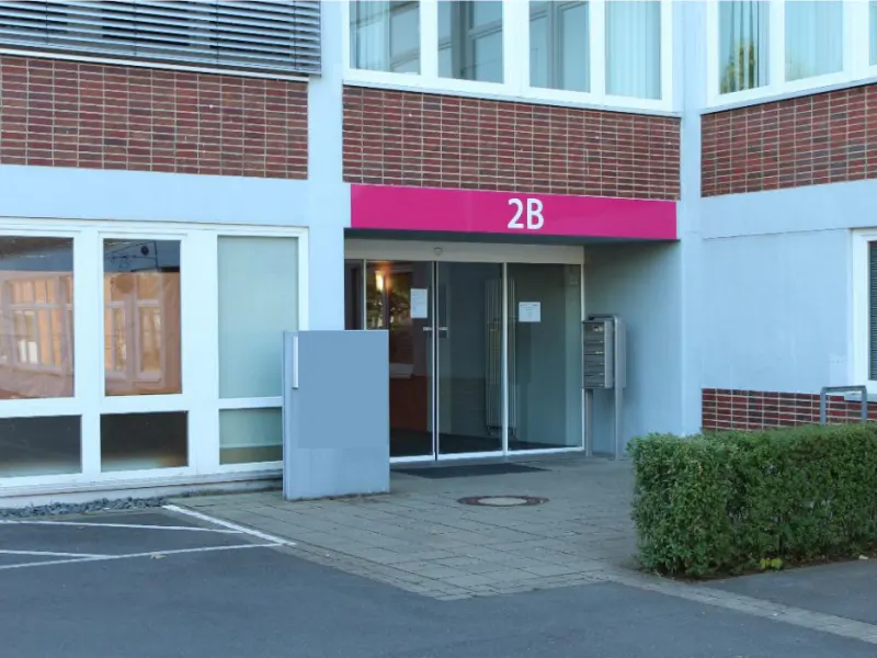 Exterior view of a building with a pink sign reading "2B" above a glass door entrance. Windows are on the left, and a mailbox is on the right.