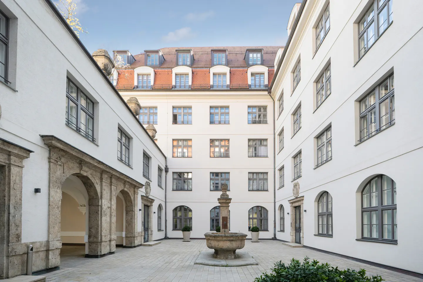 Courtyard view of a white building with a stone fountain in the center. Arched doorways and windows are visible.