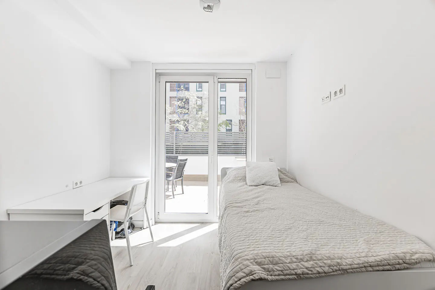 Bright, all-white bedroom with a single bed, desk, and sliding glass doors to a balcony with outdoor furniture.