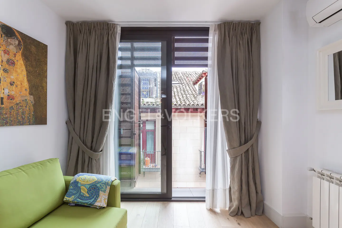 Living room with a green sofa, a Klimt painting, and a glass door to a balcony with a view of a building.