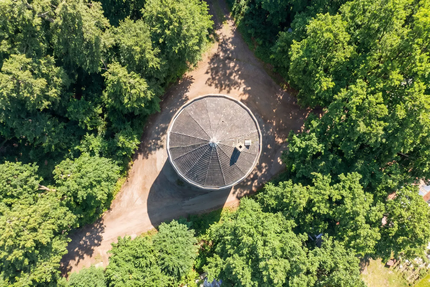 Aerial view of a round, gray-roofed building surrounded by green trees and dirt paths.