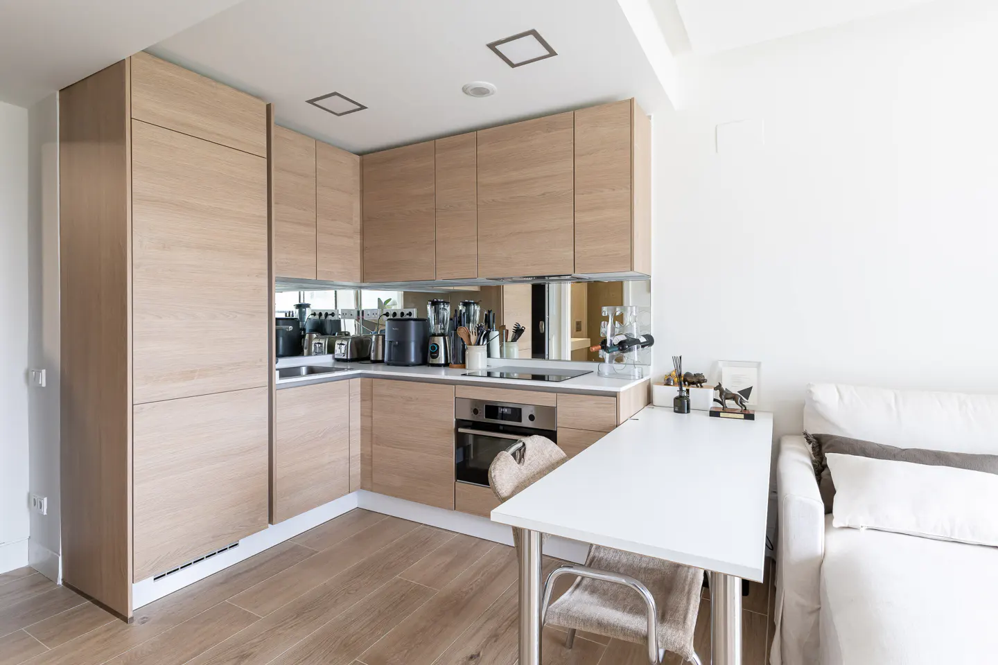A modern kitchen with light wood cabinets, mirrored backsplash, white countertop, oven, and a white table with two chairs next to a white sofa.