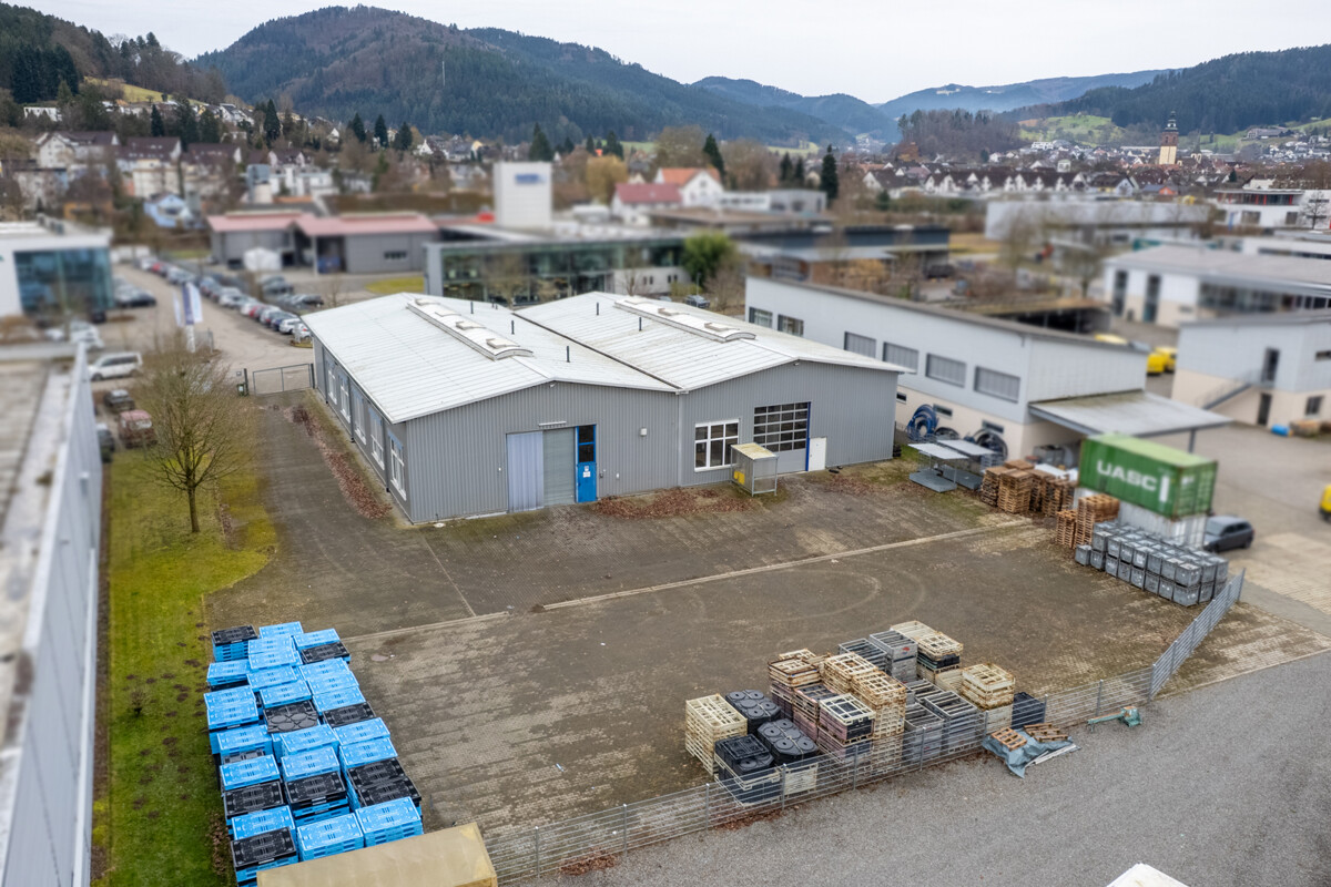 Aerial view of a gray industrial building with a white roof, surrounded by pallets and a mountain backdrop.