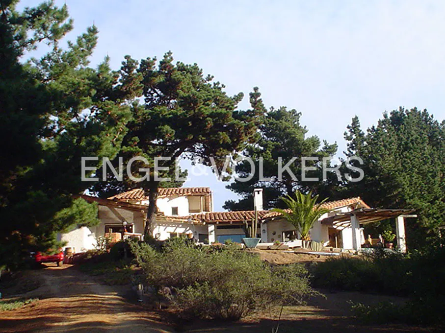 Exterior view of a white house with a red tile roof, surrounded by green trees and bushes. The Engel & Volkers logo is visible.