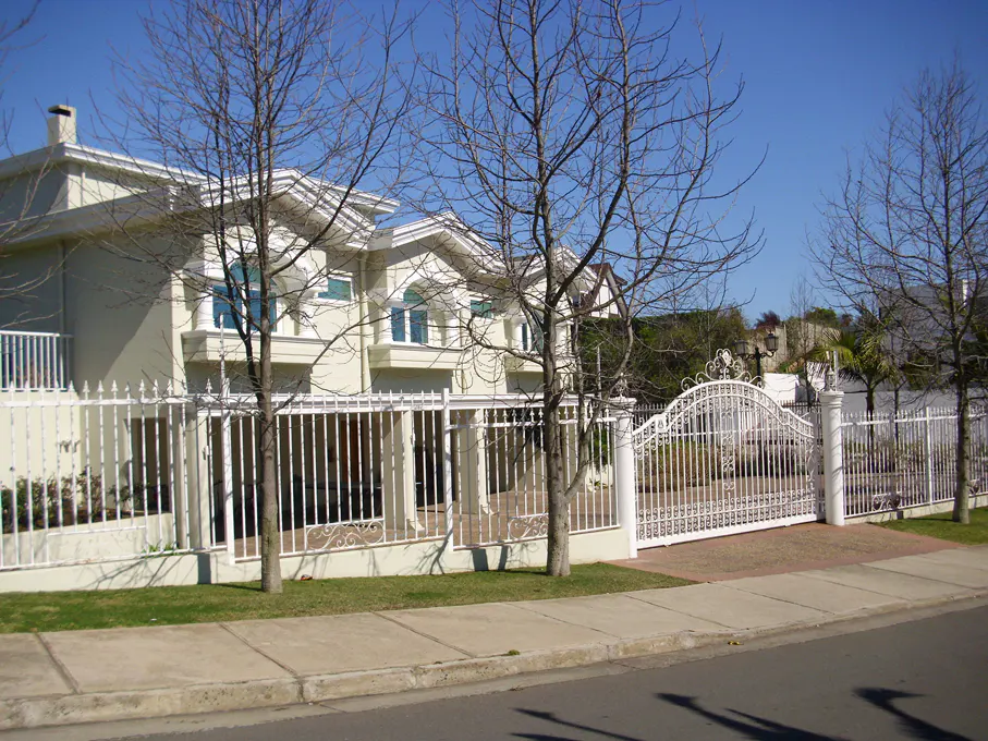 A two-story beige house with a white fence and gate, viewed from the street on a sunny day.
