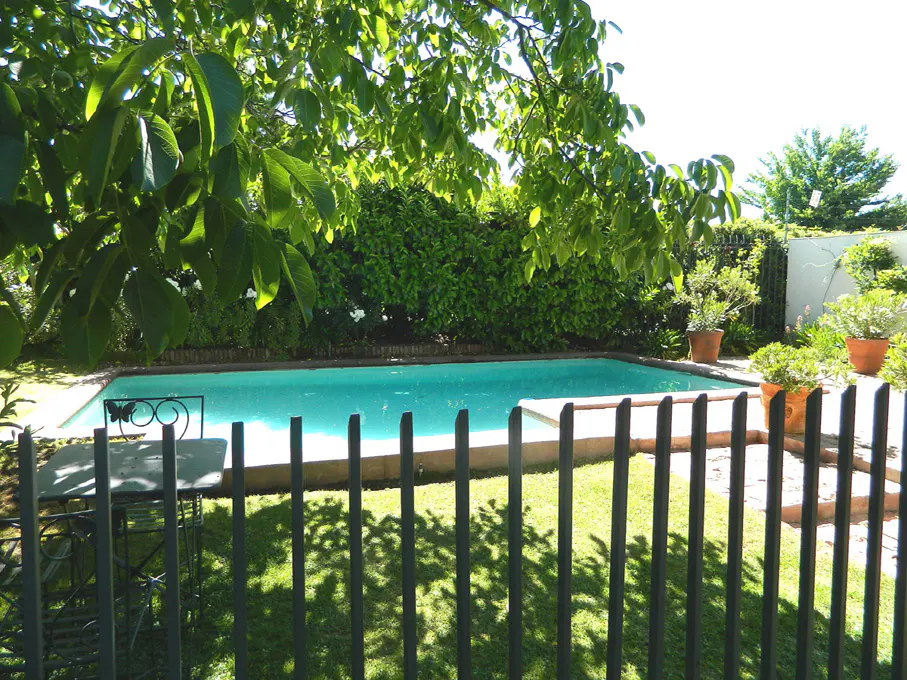 A backyard swimming pool with turquoise water is surrounded by green trees and a black metal fence.