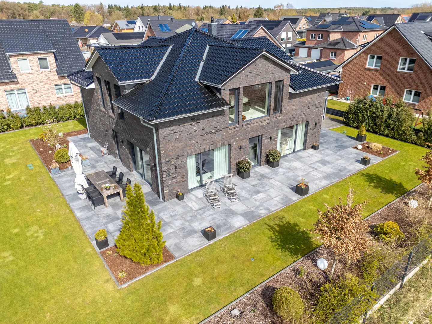 Aerial view of a modern, two-story brick house with a black roof, a stone patio, and a green lawn.
