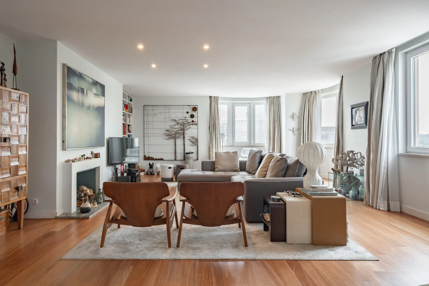 Bright living room with wood floors, a gray sectional sofa, and two brown leather chairs on a white rug. Artwork adorns the walls.
