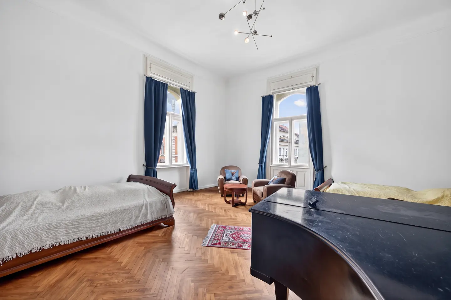 Bright room with hardwood floors, white walls, and blue curtains. Features a bed, chairs, a table, a rug, and a black piano.