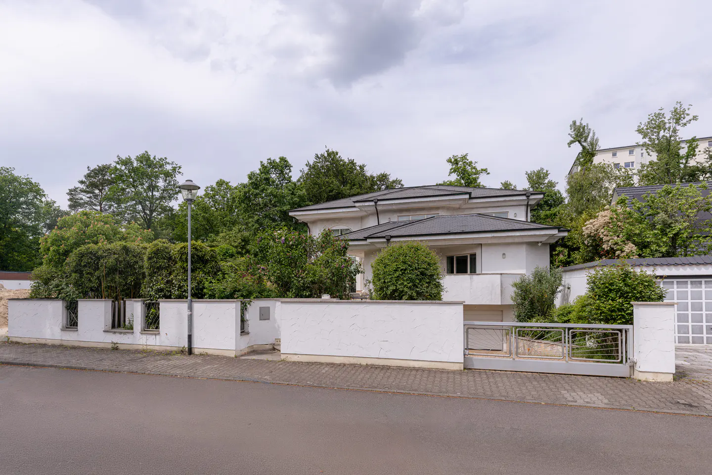 A modern white two-story house with a dark roof, surrounded by lush green trees and a white wall with a metal gate.