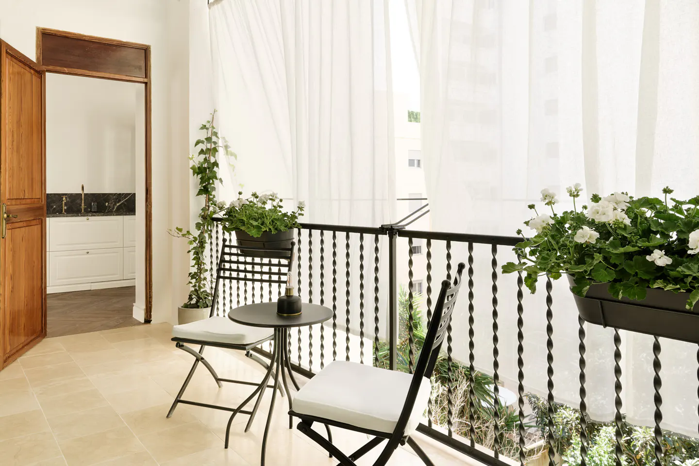 A bright balcony with black metal furniture, white cushions, and flower boxes with white flowers. Sheer white curtains hang in the background.
