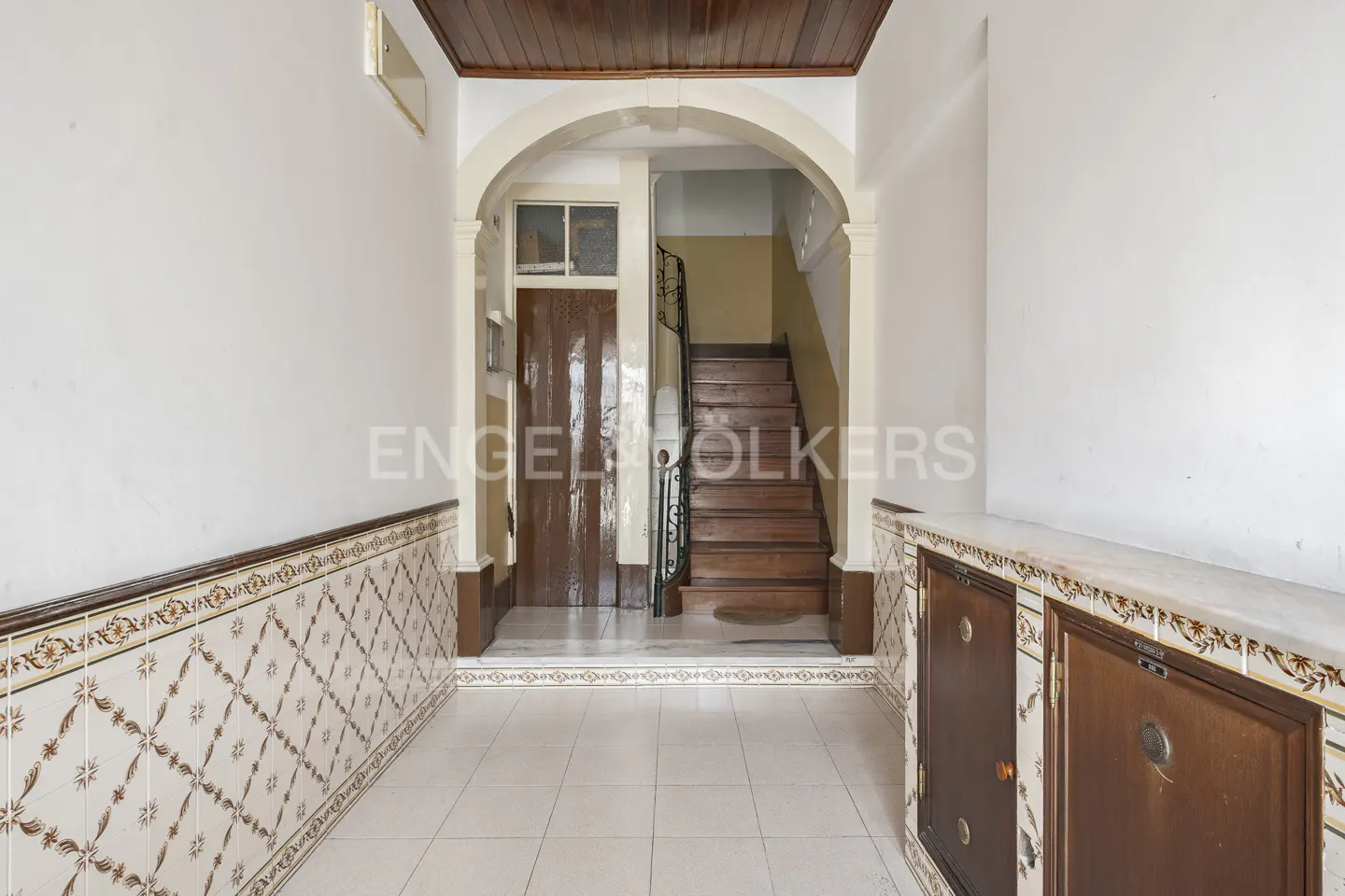 Hallway with white walls, tiled wainscoting, and a wooden ceiling. An arched doorway leads to a staircase and a brown door.
