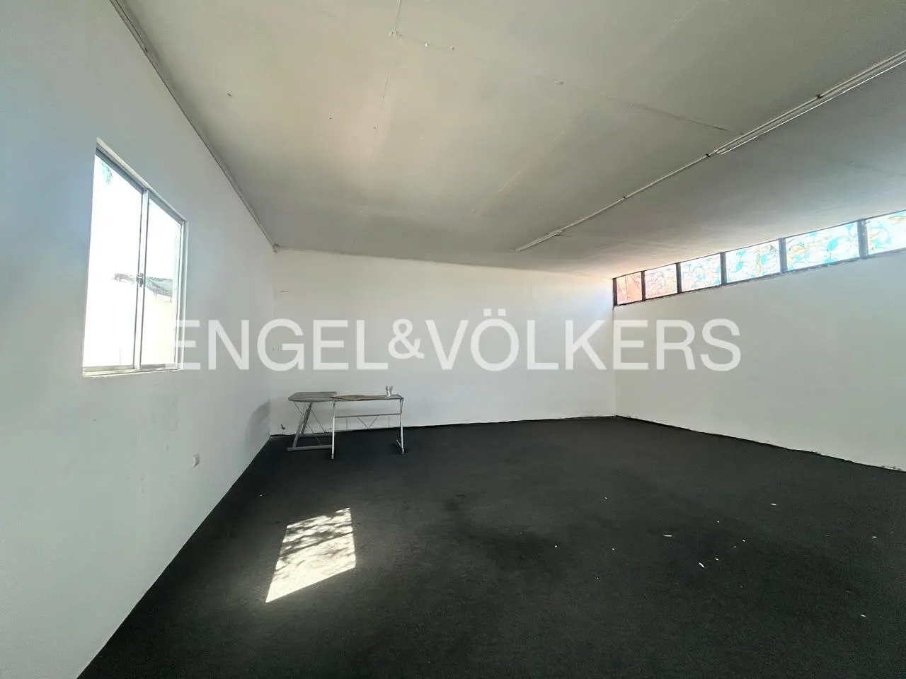 Empty white room with black carpet, a metal table, and windows letting in light. Engel & Völkers logo is superimposed.