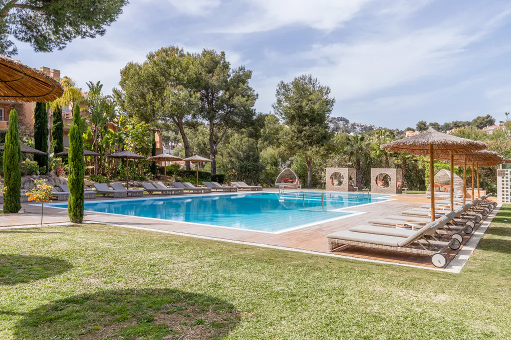 Outdoor pool with lounge chairs and straw umbrellas on a sunny day. Trees and buildings in the background.