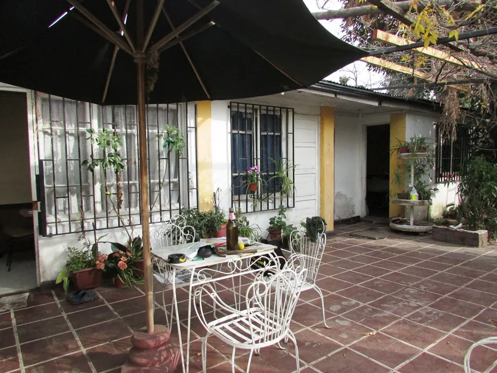 Outdoor patio with white metal table and chairs under a black umbrella. Potted plants line the white and yellow house with barred windows.