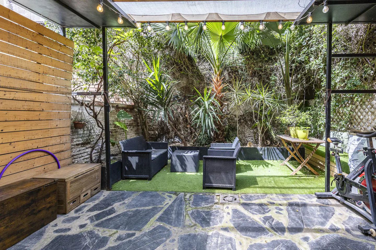 Outdoor patio with stone floor, wood fence, and lush greenery. Black wicker furniture sits on artificial turf. A wooden table and exercise bike are also present.