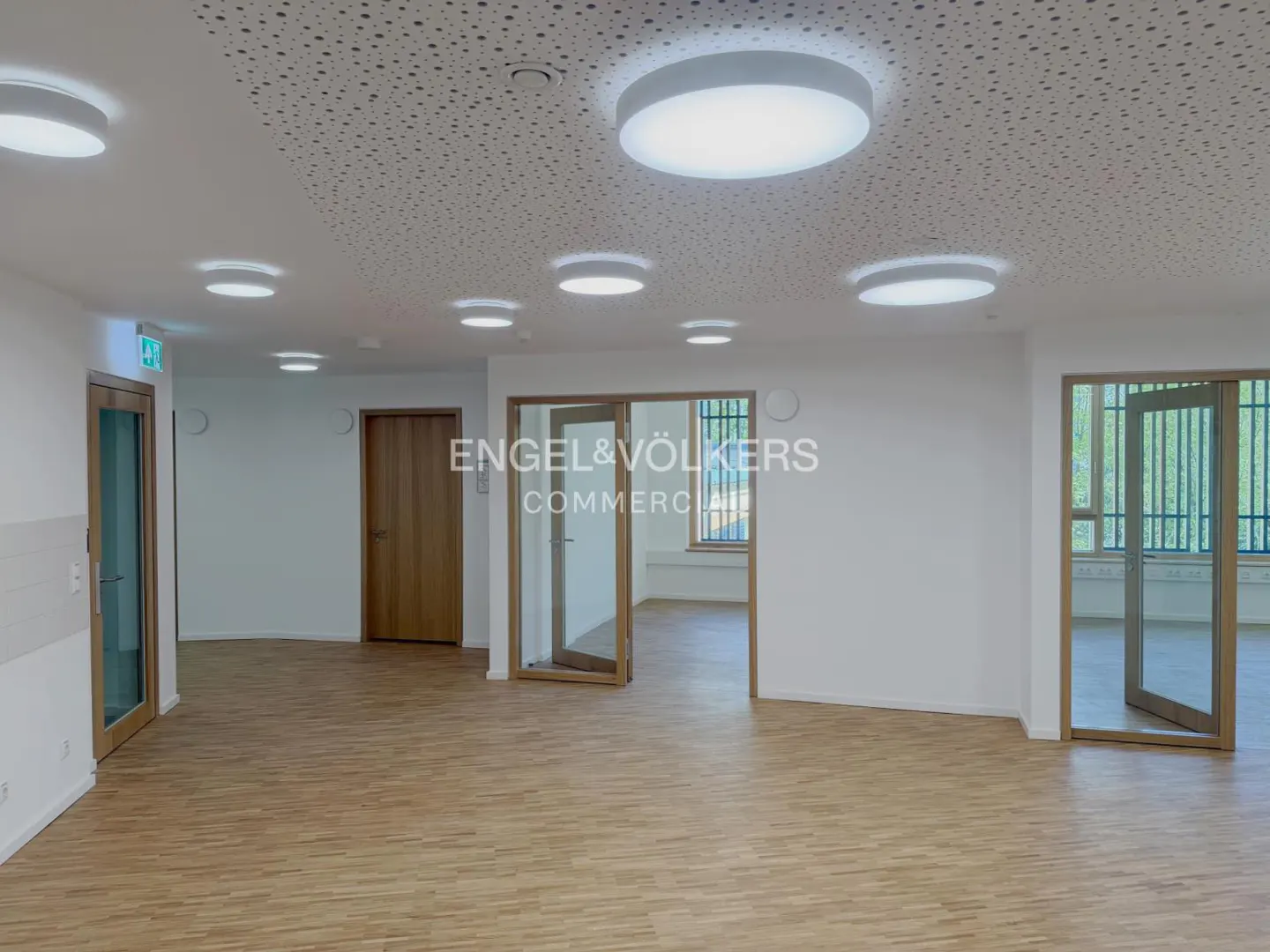 Empty room with wood floor, white walls, and round ceiling lights. Doors and windows are visible.