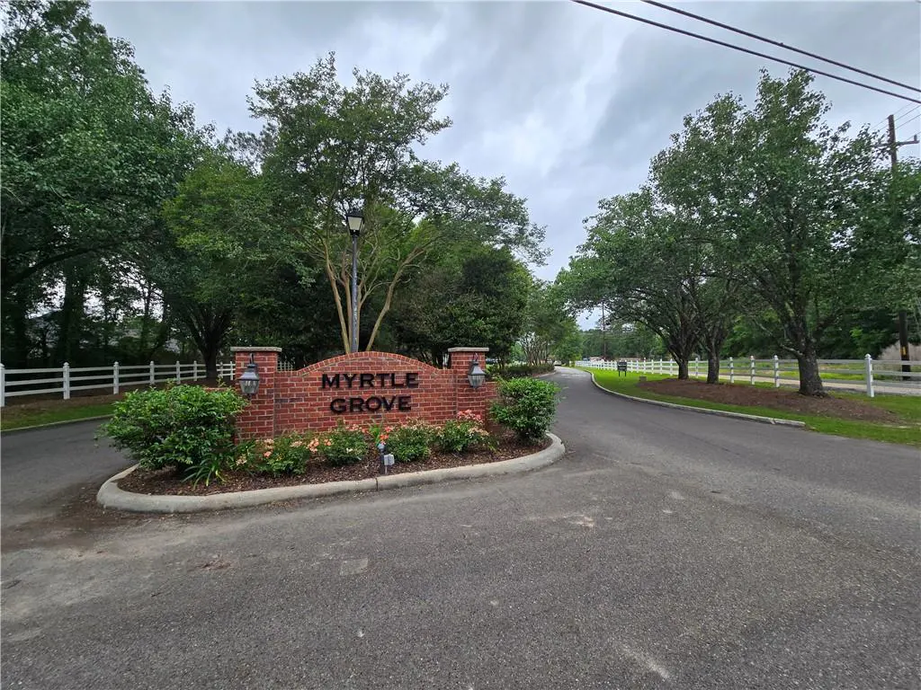 Entrance to Myrtle Grove with a brick sign, green bushes, and trees under a cloudy sky. A white fence lines the road.