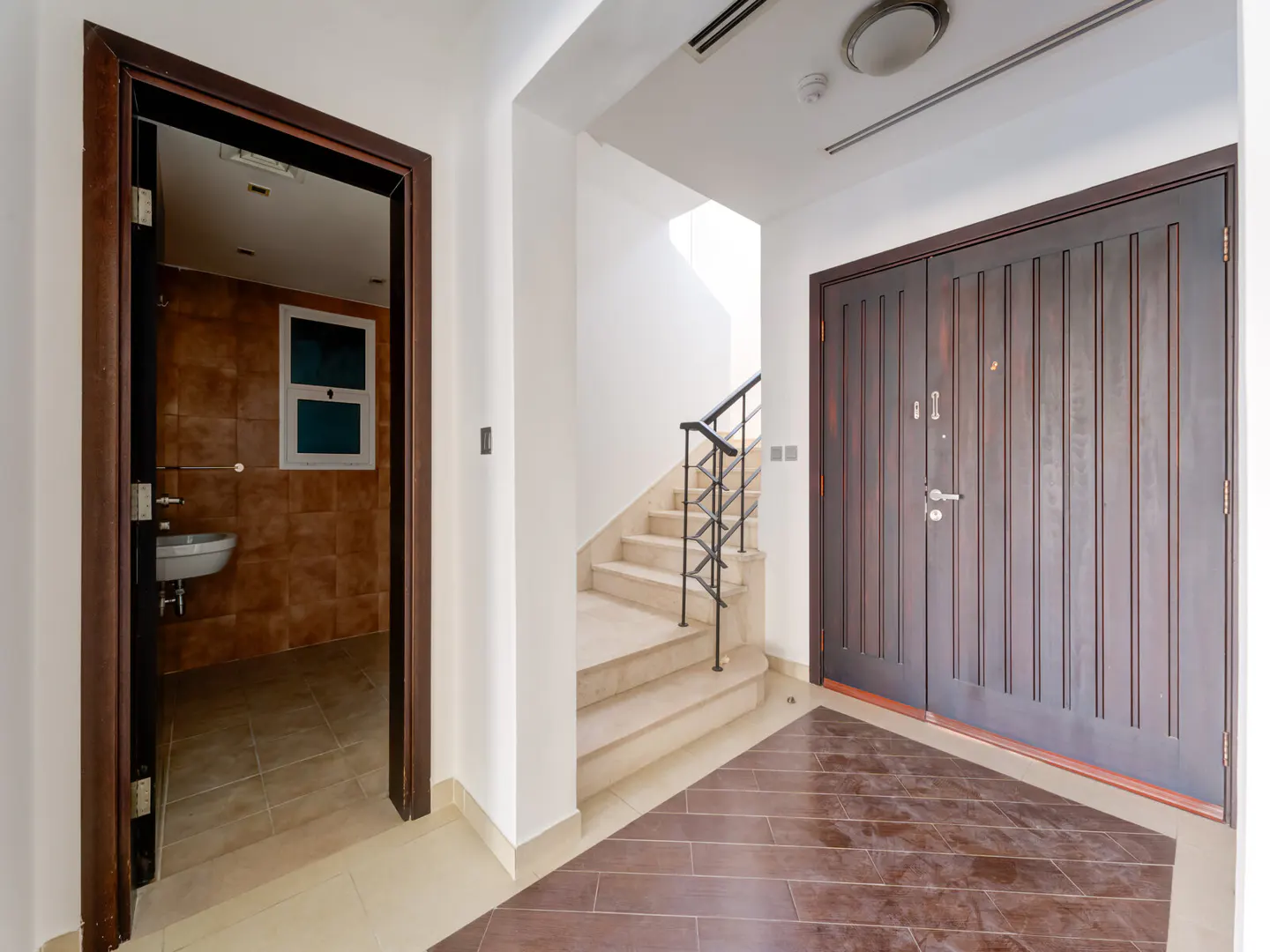 Entryway with dark wood double doors, stairs with black railing, and open bathroom with brown tile.