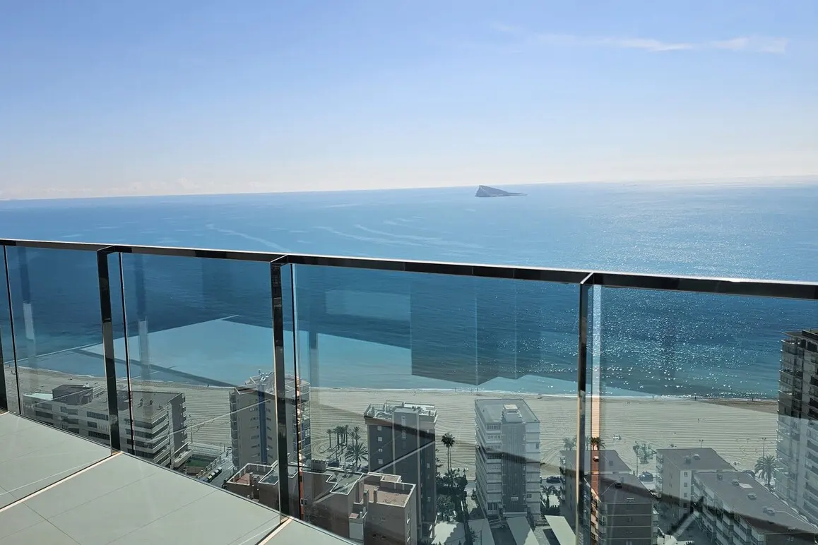 View from a high-rise balcony with glass railings overlooking a beach, ocean, and distant island under a clear blue sky.