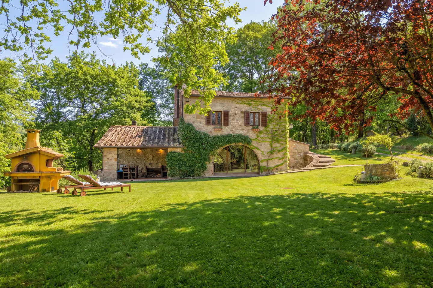 Stone house with ivy, brown shutters, and tile roof. Green lawn, trees, and outdoor pizza oven.