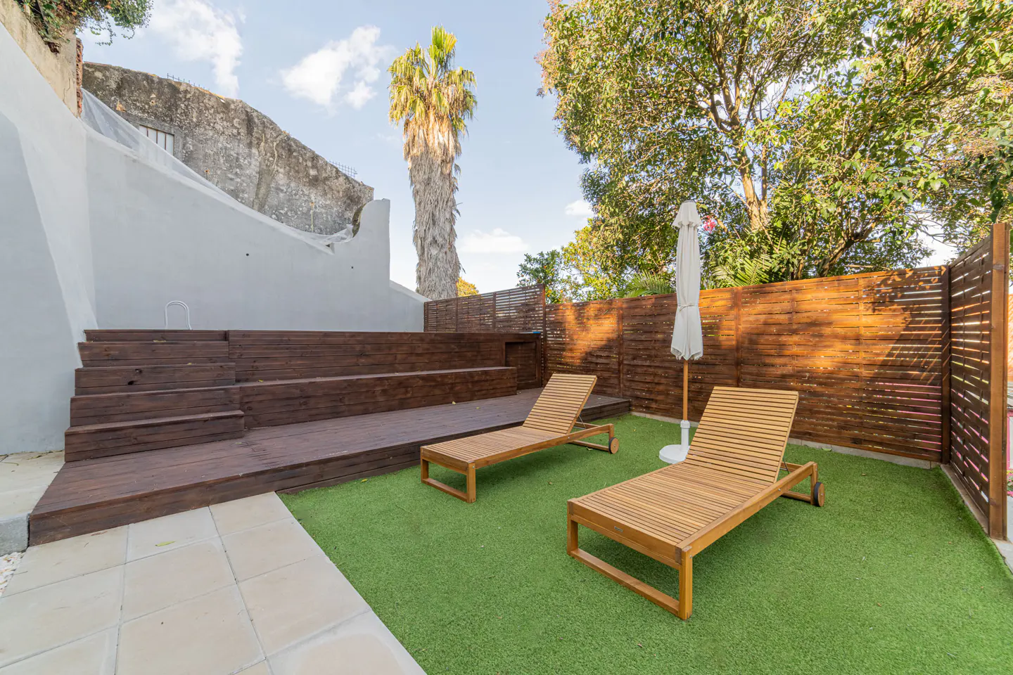 Outdoor patio with two wooden lounge chairs on green turf, a wooden deck with steps, and a white umbrella.