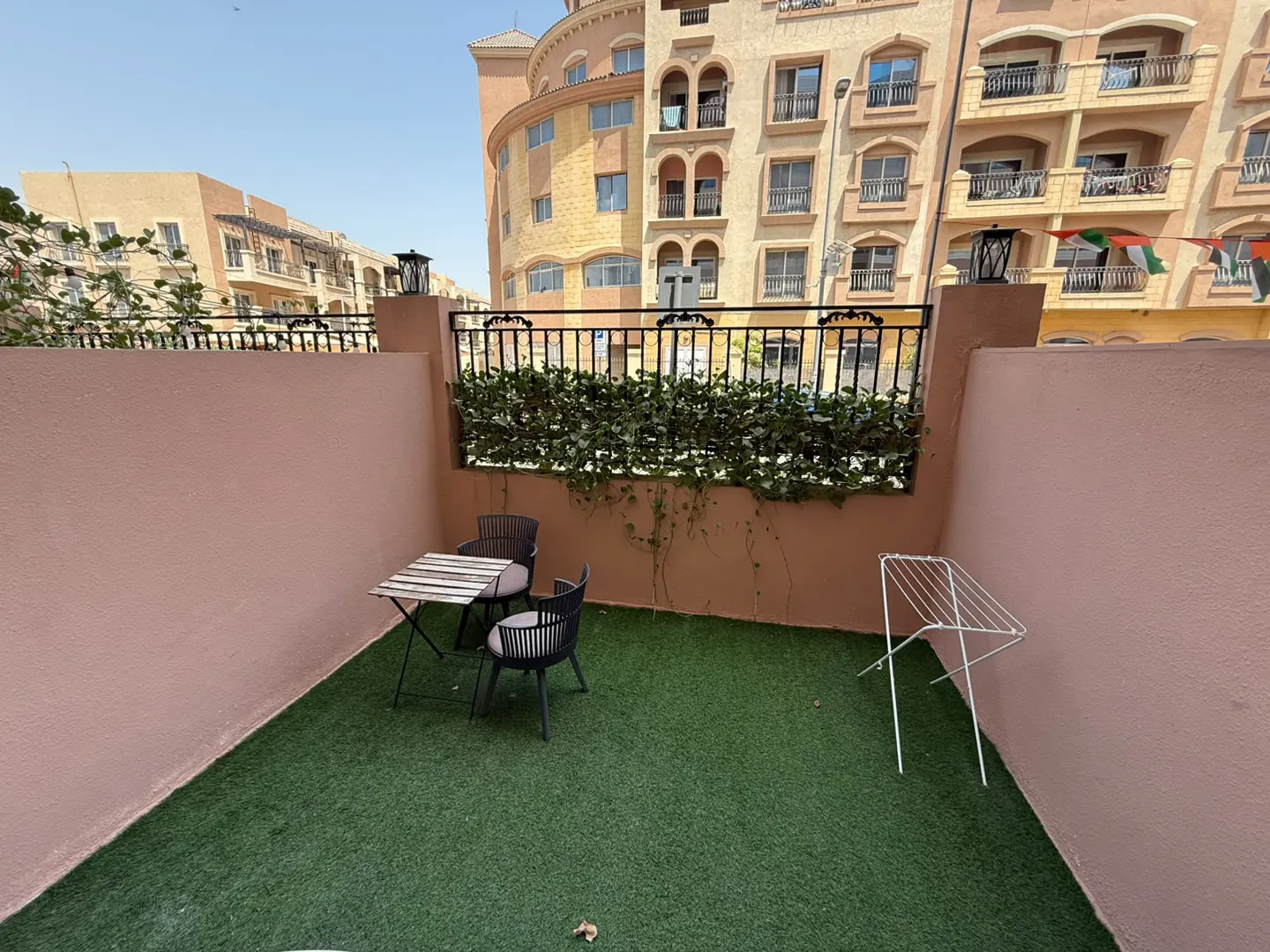 A small outdoor patio with artificial grass, a table and two chairs, and a clothes drying rack. A black iron fence with greenery sits atop a pink wall. Buildings are in the background.