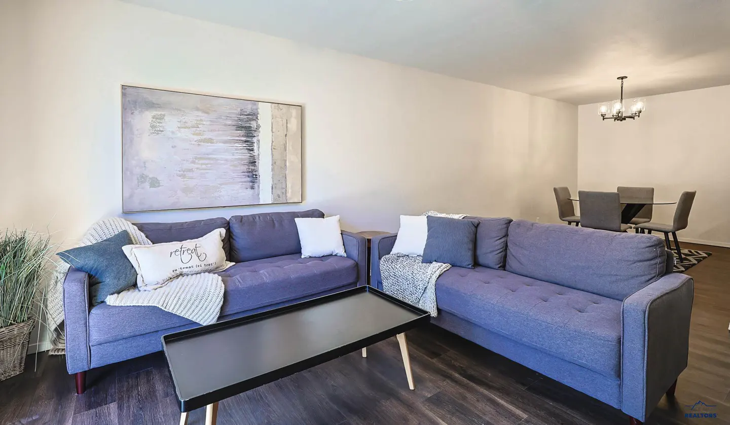 Living room with two blue sofas, a black coffee table, and a dining area with a glass table and gray chairs. An abstract painting hangs on the wall.