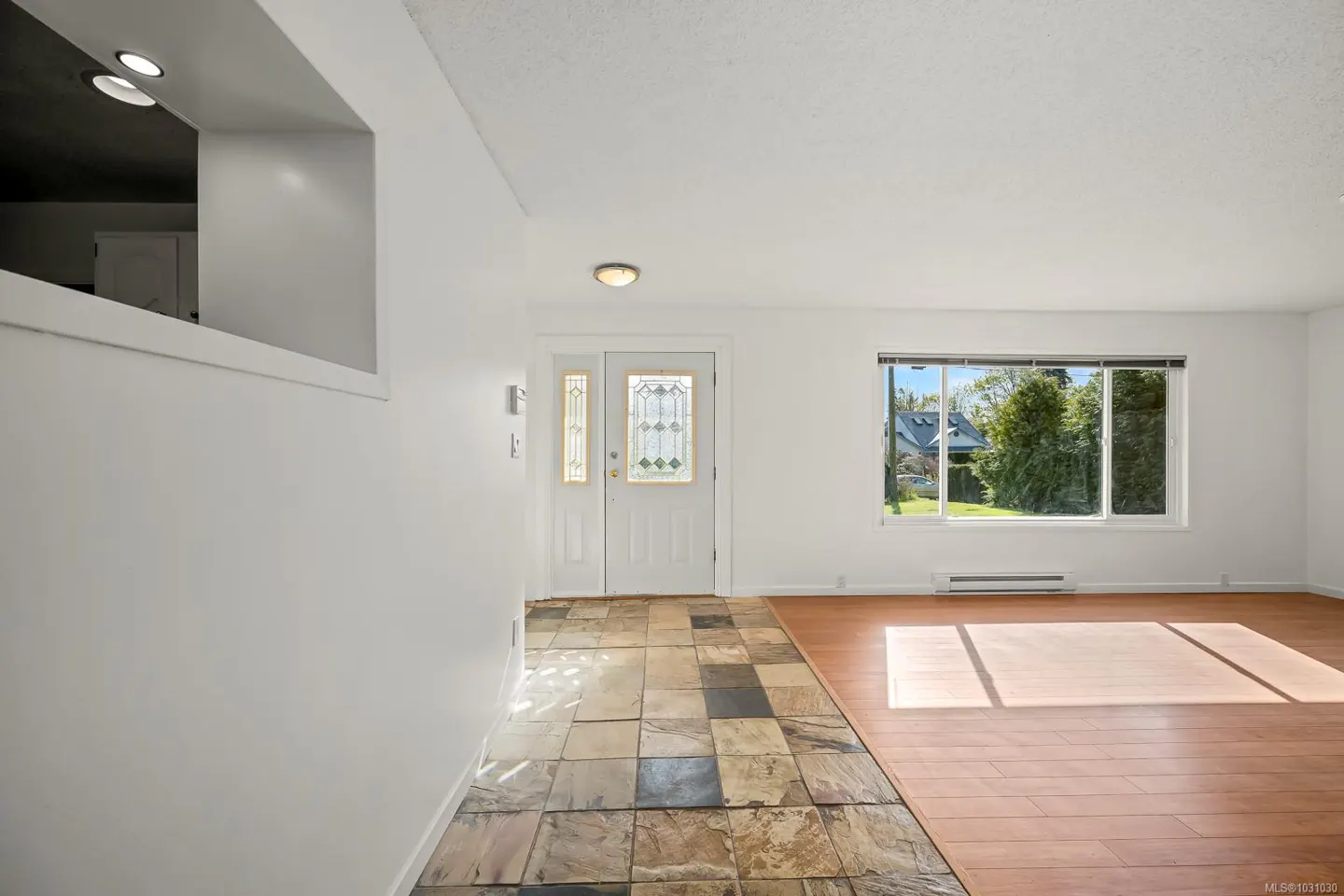 Bright entryway with white walls, stone tile floor transitioning to wood, and a front door with decorative glass. A large window offers a view of greenery.