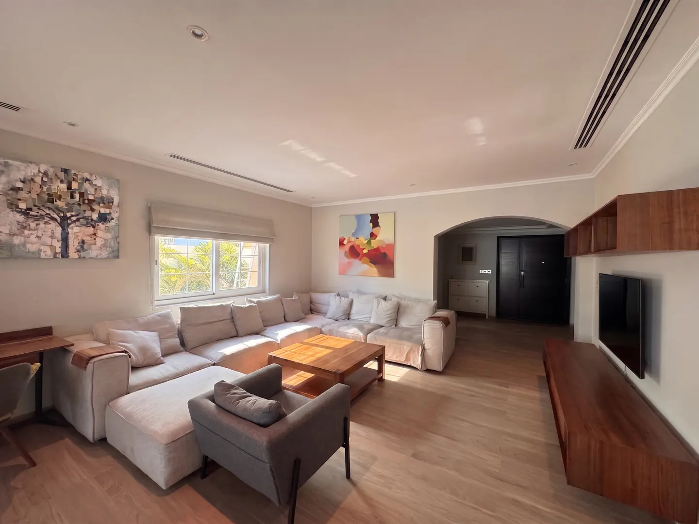 Bright living room with a large beige sectional sofa, wooden coffee table, and gray armchair. Artwork adorns the walls, and natural light streams through the window.