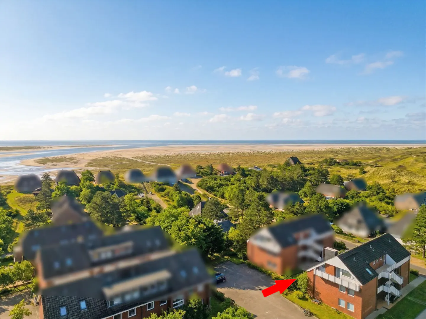 Aerial view of a red brick apartment building near a beach, under a blue sky with white clouds. An arrow points to the building.