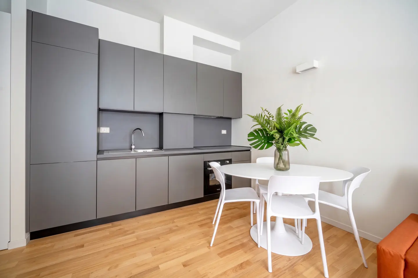 A modern kitchen with gray cabinets, a white round table, and four white chairs on a wooden floor. A vase of green plants sits on the table.