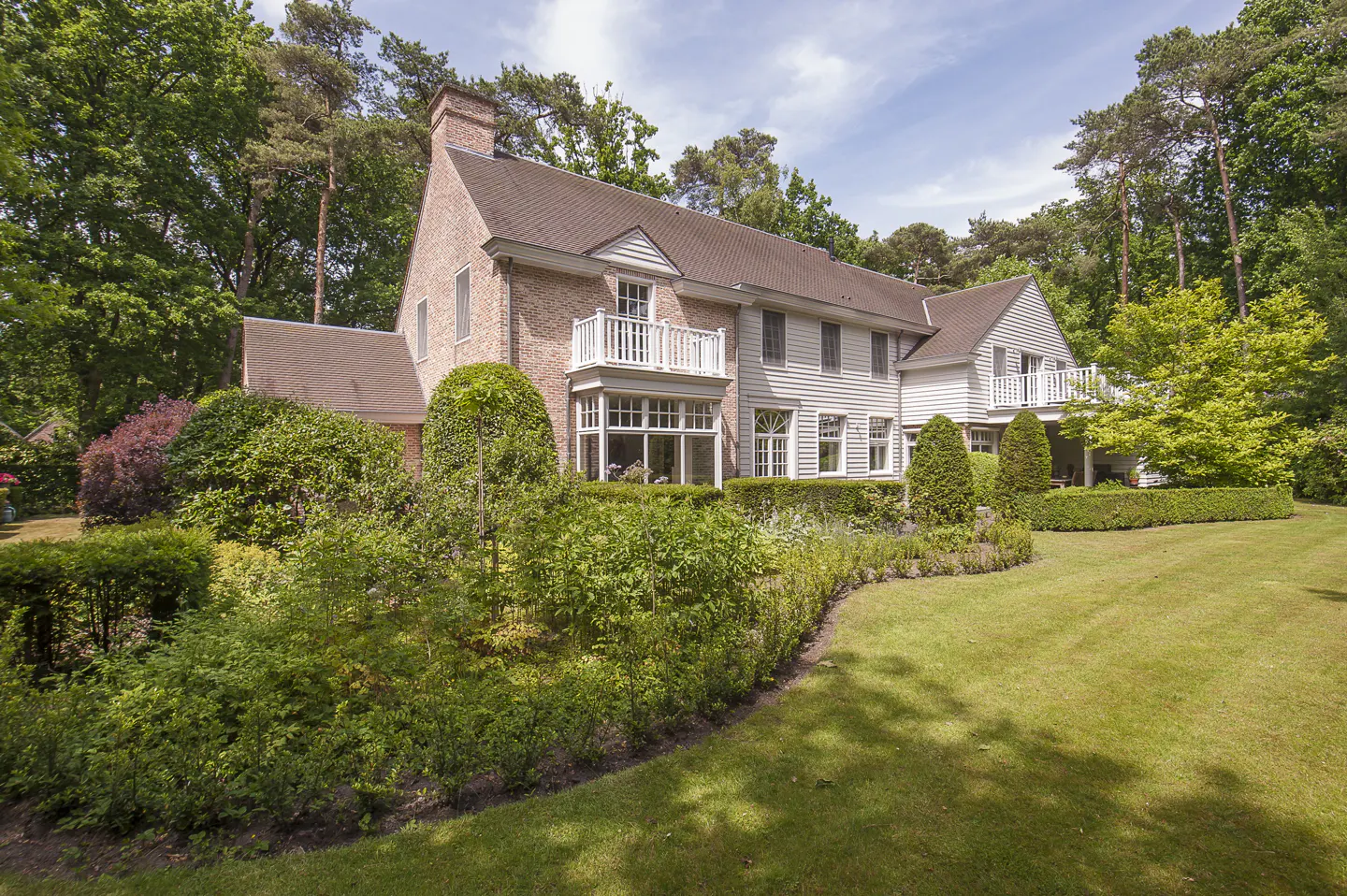 Exterior view of a large two-story house with a brick and white wood facade, balconies, and a manicured lawn surrounded by lush green trees.