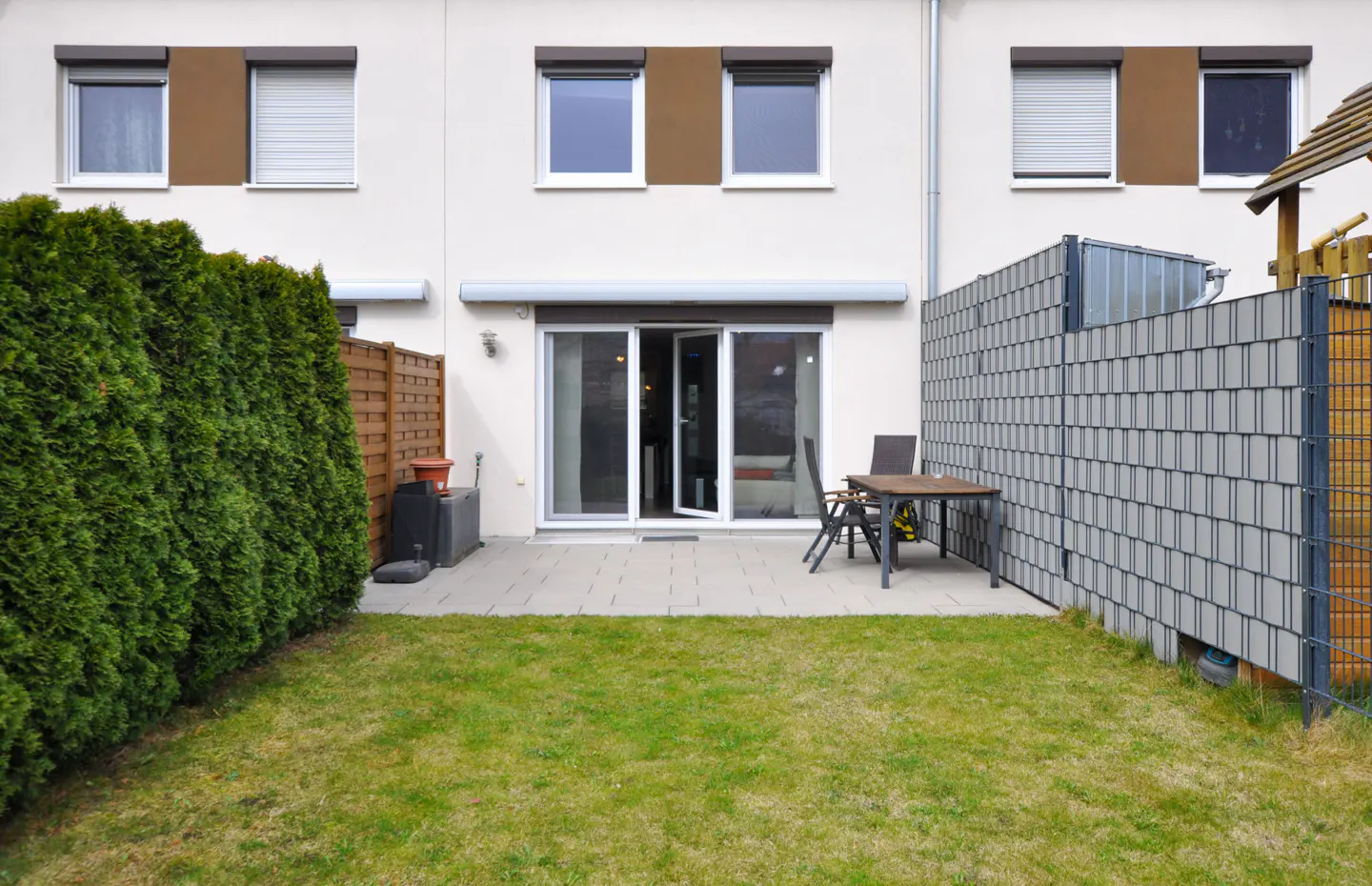 Small backyard with a paved patio, dining set, green hedge, and grey privacy fence against a white townhouse exterior.