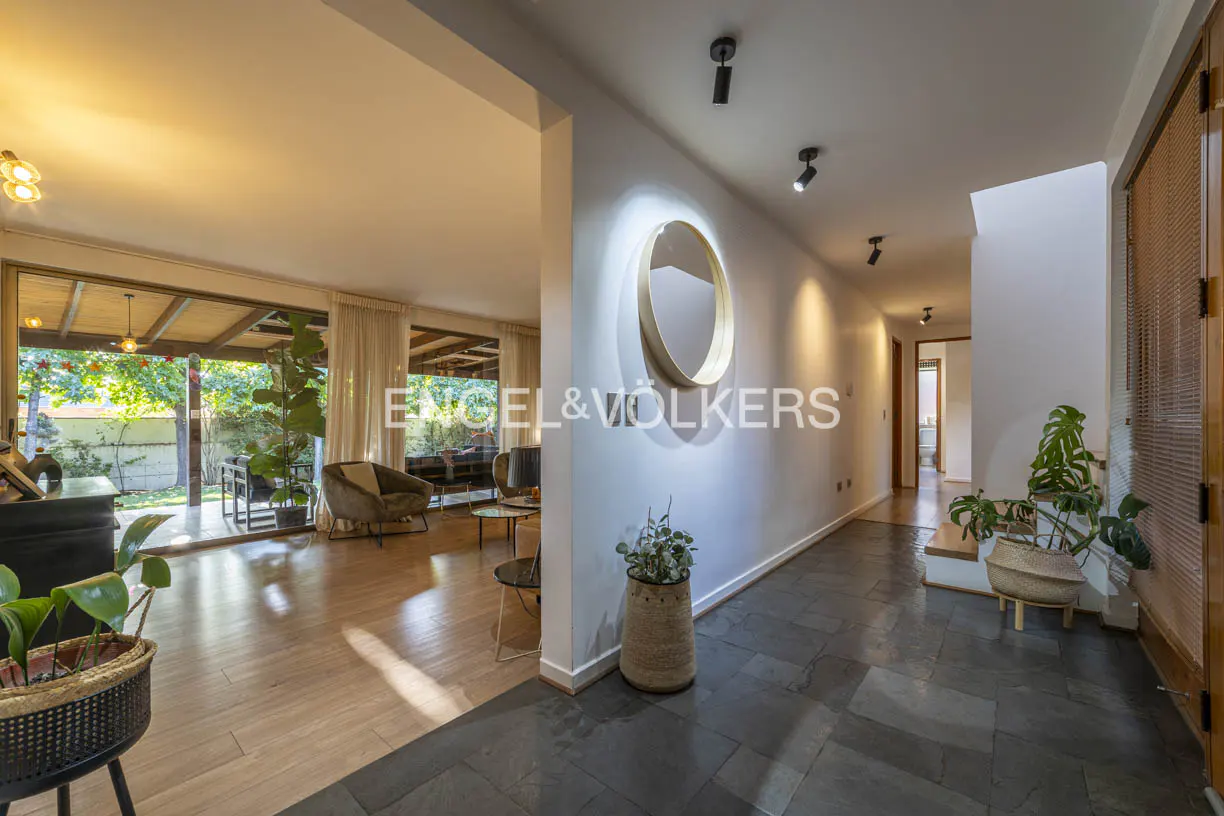 A bright home interior with wood floors, a round mirror, and plants in woven baskets. A living room is visible through a wide doorway.