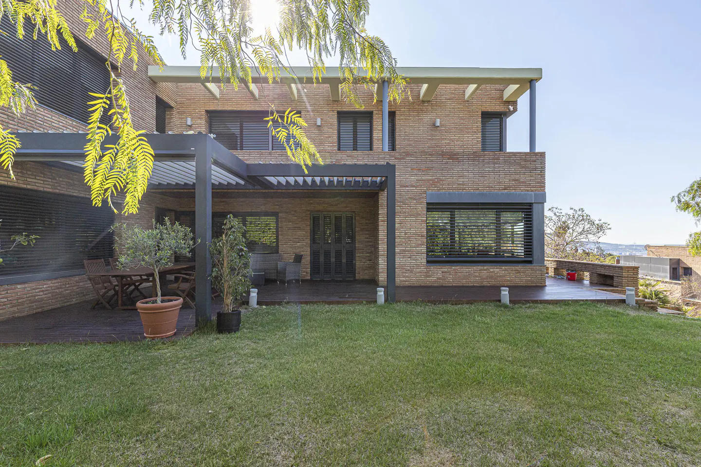 Two-story brick house with a green lawn, a wooden deck, and a pergola. The house has black window shutters and a balcony.