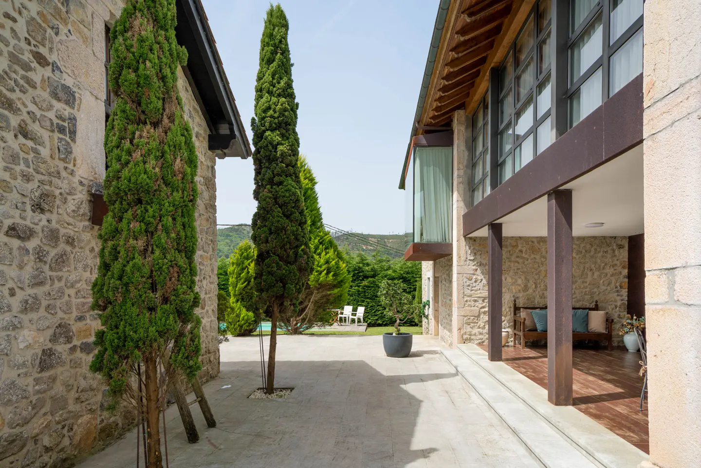 Exterior view of a stone house with tall cypress trees, a patio, and a glimpse of a pool and lawn in the background.