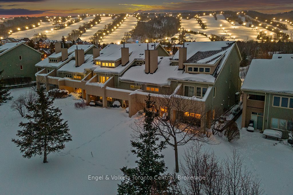 Snow-covered townhomes at dusk with a brightly lit ski hill in the background under a sunset sky. [AI-generated text]