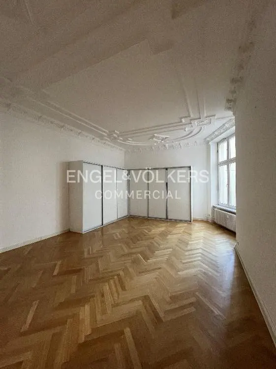 Empty room with herringbone wood floors, white walls, and a large window. Engel & Völkers Commercial logo on a white wall.