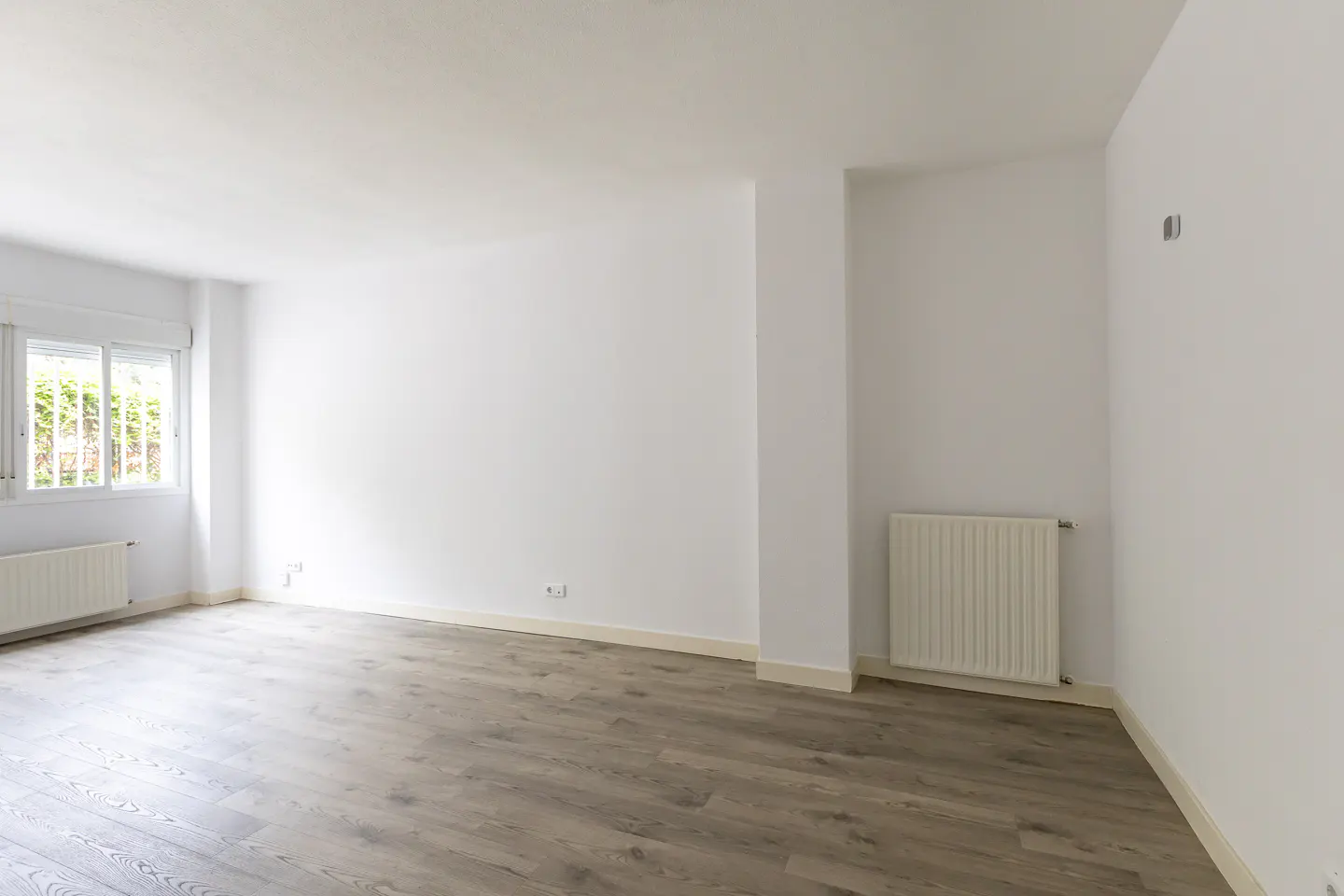 Bright, empty room with gray wood floors, white walls, and a window with green foliage outside. A white radiator sits under the window.