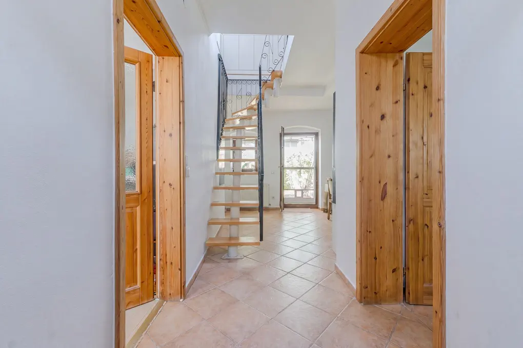 Hallway with white walls, terracotta tile floor, and wooden doors. A spiral staircase with black railing leads to the upper floor.