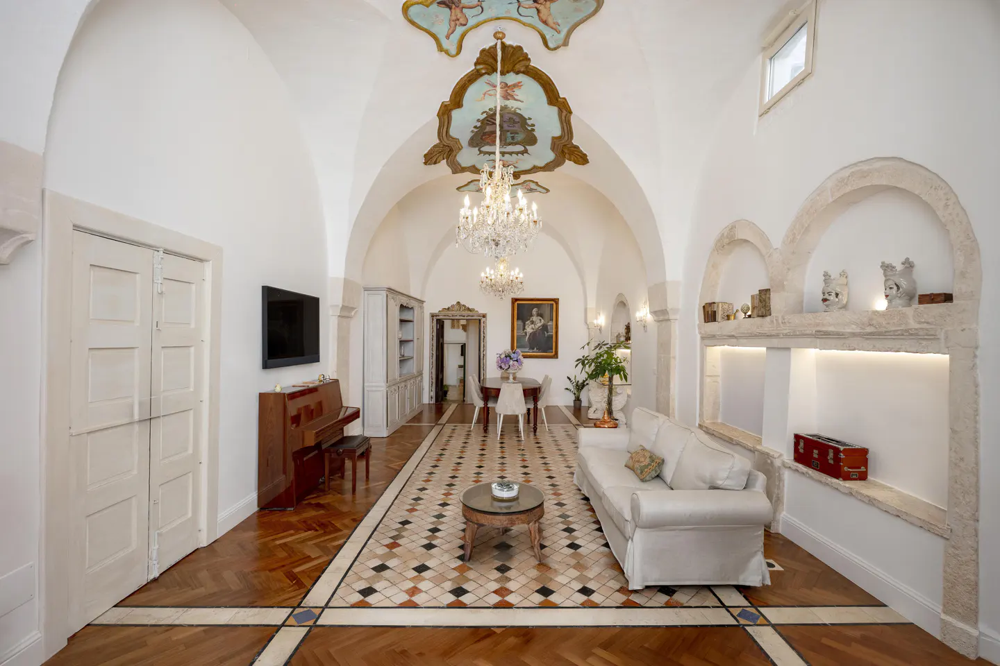 A bright, elegant living room with arched ceilings, a chandelier, and a white sofa. A piano sits near a TV, and a dining area is visible in the background.
